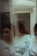 A person with curly hair is partially obscured by a reflective surface, likely a mirror, creating multiple blurred reflections. The person is wearing a light-colored, long-sleeved shirt and appears contemplative.