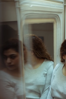 A person with curly hair is partially obscured by a reflective surface, likely a mirror, creating multiple blurred reflections. The person is wearing a light-colored, long-sleeved shirt and appears contemplative.