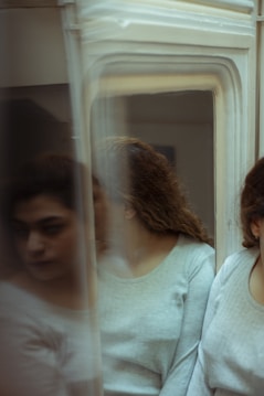 A person with curly hair is partially obscured by a reflective surface, likely a mirror, creating multiple blurred reflections. The person is wearing a light-colored, long-sleeved shirt and appears contemplative.