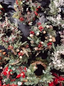 A collection of festive holiday wreaths featuring a variety of greenery, including pine and holly leaves. Accents include red berries, pine cones, and small decorative ornaments. Some wreaths also feature snow-like frosted tips and bows. The wreaths are arranged against a dark, wooden background, enhancing their vibrant colors.