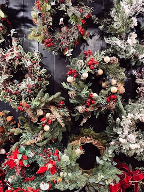 A collection of festive holiday wreaths featuring a variety of greenery, including pine and holly leaves. Accents include red berries, pine cones, and small decorative ornaments. Some wreaths also feature snow-like frosted tips and bows. The wreaths are arranged against a dark, wooden background, enhancing their vibrant colors.