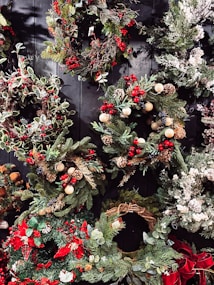 A collection of festive holiday wreaths featuring a variety of greenery, including pine and holly leaves. Accents include red berries, pine cones, and small decorative ornaments. Some wreaths also feature snow-like frosted tips and bows. The wreaths are arranged against a dark, wooden background, enhancing their vibrant colors.