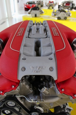 A detailed close-up of a V12 engine with a red and silver color scheme, prominently displaying the brand name. In the background, several engines are displayed on a yellow surface, with a red sports car partially visible.