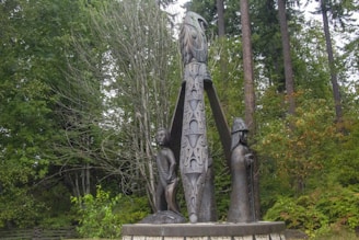 A wooden totem pole surrounded by trees, featuring intricately carved figures, including a bird and two human-like statues. The setting is lush with greenery, indicating a forested area.