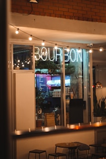 A cozy café interior is visible through the window, illuminated by a neon sign displaying 'Bourbon.' The space is warmly lit, with decorative string lights hanging outside. Tables and stools are positioned near the window, and plants add a touch of greenery inside the café. A menu board is seen in the background, along with various items on shelves.