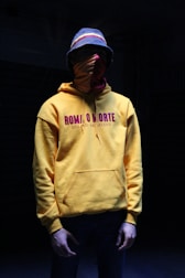 A person is wearing a bright yellow hoodie with the text 'ROMA O MORTE' printed on it, accompanied by Roman numerals. They have a multicolored bucket hat and a red cloth covering their face. The setting is dark, creating a shadowy ambiance.