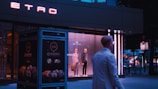 A storefront window displays two mannequins dressed in stylish clothing. The neon sign of the brand 'ETRO' glows above, while a person in a white coat walks past. The setting appears to be urban with dim evening lighting.