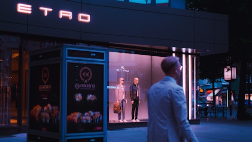 A storefront window displays two mannequins dressed in stylish clothing. The neon sign of the brand 'ETRO' glows above, while a person in a white coat walks past. The setting appears to be urban with dim evening lighting.