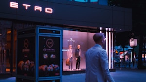 A storefront window displays two mannequins dressed in stylish clothing. The neon sign of the brand 'ETRO' glows above, while a person in a white coat walks past. The setting appears to be urban with dim evening lighting.