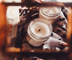 Two glass jars with white candles inside are placed on a floral patterned fabric. The scene is softly lit, creating a cozy and warm atmosphere. Additionally, there are some illustrated cards or stamps visible, adding to the aesthetic appeal.