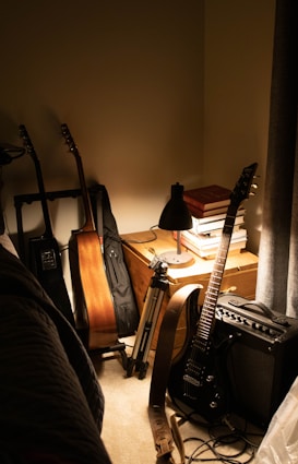 In a dimly lit room, a wooden guitar stands upright next to a black electric guitar leaning against an amplifier. Nearby, a camera tripod is positioned next to a small desk with a black lamp and stacks of books. The scene is organized yet cozy, with natural wood tones and soft lighting adding to the ambiance.