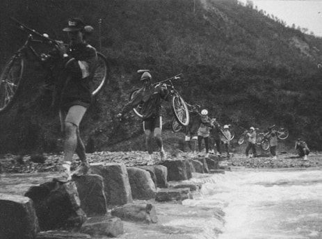 A group of individuals carrying bicycles walk across a series of large stones in a river. The setting appears to be outdoors in a rugged, mountainous area. The people are dressed in outdoor gear and helmets, suggesting a cycling expedition. The atmosphere is adventurous, with a focus on teamwork and resilience as they navigate the river crossing.