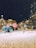 A festive winter scene in a city square with snow covering the ground. Large, colorful candy decorations and artificial figures resembling elves and mice are arranged around a beautifully lit and ornamented Christmas tree. Buildings in the background are adorned with bright, warm lights while snow is gently falling from the night sky.