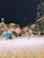 A festive winter scene in a city square with snow covering the ground. Large, colorful candy decorations and artificial figures resembling elves and mice are arranged around a beautifully lit and ornamented Christmas tree. Buildings in the background are adorned with bright, warm lights while snow is gently falling from the night sky.