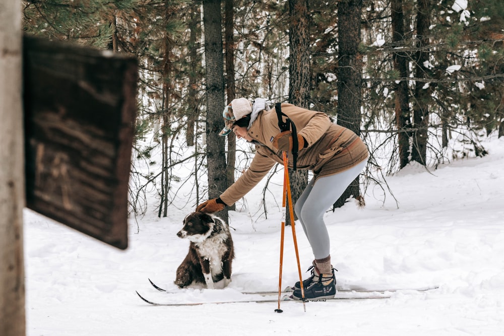 man in brown jacket and black pants holding black and white dog on snow covered ground