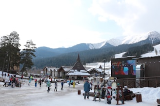 A bustling ski resort area with several people walking and skiing near wooden buildings. There is a large screen displaying a video, and mountains covered in snow and pine trees are visible in the background. Ski equipment is stored in racks to the right.