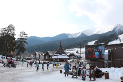 A bustling ski resort area with several people walking and skiing near wooden buildings. There is a large screen displaying a video, and mountains covered in snow and pine trees are visible in the background. Ski equipment is stored in racks to the right.