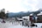 A bustling ski resort area with several people walking and skiing near wooden buildings. There is a large screen displaying a video, and mountains covered in snow and pine trees are visible in the background. Ski equipment is stored in racks to the right.