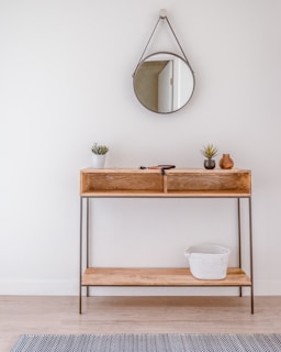 A minimalist wooden console table against a white wall with a round mirror hanging above it. The table holds small potted plants and decorative vases. A woven basket is placed on the lower shelf. Light wooden flooring and a subtle patterned rug complete the setting.