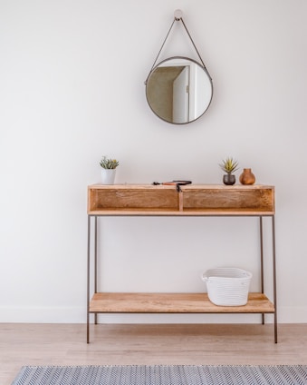 A minimalist wooden console table against a white wall with a round mirror hanging above it. The table holds small potted plants and decorative vases. A woven basket is placed on the lower shelf. Light wooden flooring and a subtle patterned rug complete the setting.