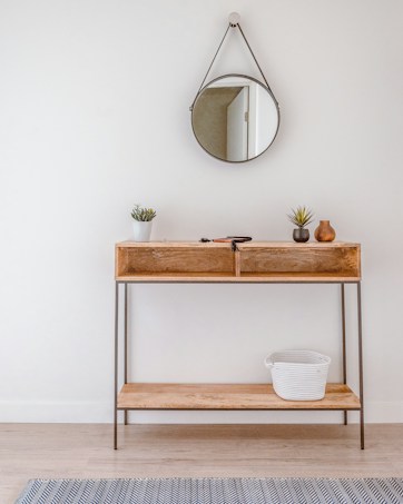A minimalist wooden console table against a white wall with a round mirror hanging above it. The table holds small potted plants and decorative vases. A woven basket is placed on the lower shelf. Light wooden flooring and a subtle patterned rug complete the setting.