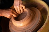 Close-up of a student carefully shaping a clay pot on a spinning wheel under expert guidance.