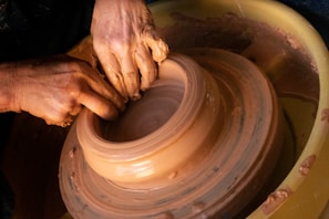 Close-up of a student carefully shaping a clay pot on a spinning wheel under expert guidance.