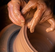 Hands covered in clay delicately shape a pot on a potter’s wheel, showcasing the intricate craft of pottery making. The brown, wet clay spins smoothly as the fingers mold it with precision.