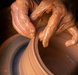 Hands covered in clay delicately shape a pot on a potter’s wheel, showcasing the intricate craft of pottery making. The brown, wet clay spins smoothly as the fingers mold it with precision.