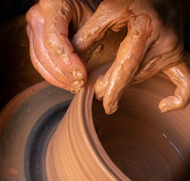 Hands covered in clay delicately shape a pot on a potter’s wheel, showcasing the intricate craft of pottery making. The brown, wet clay spins smoothly as the fingers mold it with precision.