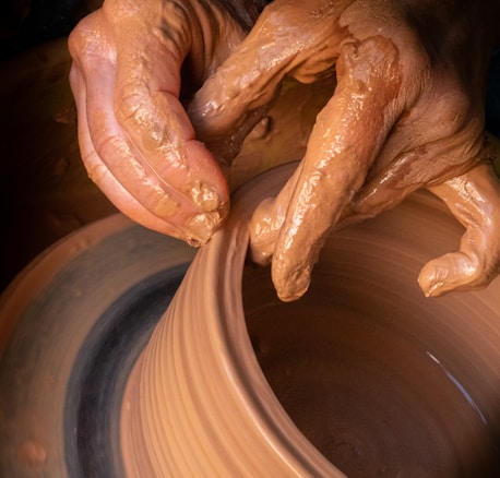 Hands covered in clay delicately shape a pot on a potter’s wheel, showcasing the intricate craft of pottery making. The brown, wet clay spins smoothly as the fingers mold it with precision.