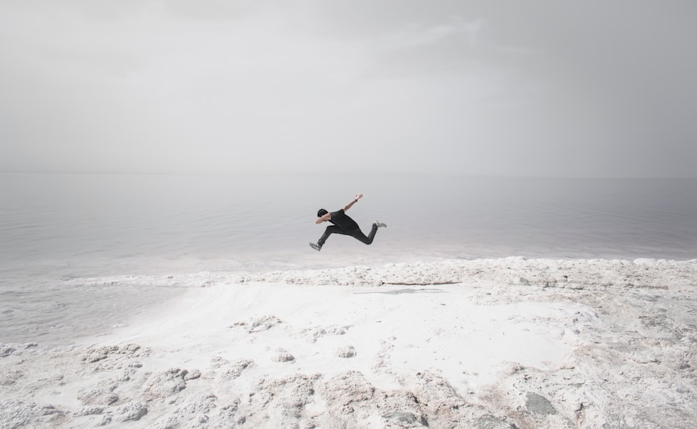 homme en chemise noire et pantalon noir sautant sur une plage de sable blanc pendant la journée