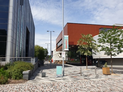 A modern urban scene featuring two large buildings, one with reflective glass panels and the other with a sleek red and white facade. Trees line a paved walkway with a patterned design. A street sign and posts are visible along with a signboard.