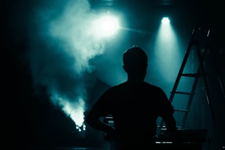 A silhouette of a person stands facing stage lights, surrounded by fog that creates a dramatic atmosphere. On the right, a ladder is visible, and the scene suggests preparation or performance in a dimly lit setting.
