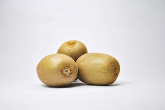 Three whole kiwi fruits arranged on a plain white background. The fruits have a brown, fuzzy skin, typical of kiwis, and are positioned in a triangular formation. The lighting is neutral, casting soft shadows beneath the fruits.