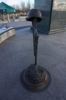A bronze sculpture depicting a military helmet set atop a rifle, with the rifle's barrel pointed downwards, embedded into a stone or rocky base. The background shows a memorial site with a large vertical stone slab and a distant pavilion structure with a green roof.