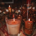Several lit candles in glass holders surrounded by a decorative arrangement, including red beads and a metallic candle holder. Soft bokeh lights create a warm and festive atmosphere.