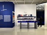 An art shop interior with a display area featuring books and items on blue tables and shelves against a white and blue background. A large wall showcases information about Kim Whanki with descriptive text and art pieces hanging on the wall. A person stands to the right, seemingly browsing on their phone.
