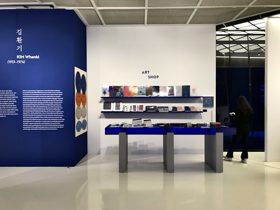 An art shop interior with a display area featuring books and items on blue tables and shelves against a white and blue background. A large wall showcases information about Kim Whanki with descriptive text and art pieces hanging on the wall. A person stands to the right, seemingly browsing on their phone.