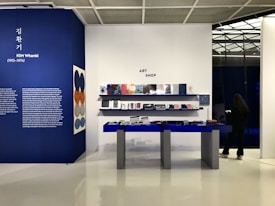 An art shop interior with a display area featuring books and items on blue tables and shelves against a white and blue background. A large wall showcases information about Kim Whanki with descriptive text and art pieces hanging on the wall. A person stands to the right, seemingly browsing on their phone.