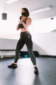 A person wearing a black sports mask and athletic attire is holding a dumbbell in a brightly lit gym with exercise mats and equipment in the background.