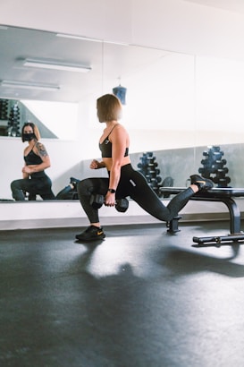 A woman is performing a Bulgarian split squat with a dumbbell in each hand in a gym setting. She is wearing athletic clothing, and her posture indicates focus and strength. A mirror reflects her image and shows a person wearing a mask in the background.