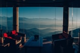 A luxurious seating area with wicker furniture and red cushions is set against a stunning backdrop of mountain ranges visible through large glass windows. The atmosphere is serene and inviting, providing a perfect blend of interior comfort with natural beauty. The misty layers of mountains stretch into the distance under a clear blue sky.
