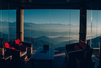 A luxurious seating area with wicker furniture and red cushions is set against a stunning backdrop of mountain ranges visible through large glass windows. The atmosphere is serene and inviting, providing a perfect blend of interior comfort with natural beauty. The misty layers of mountains stretch into the distance under a clear blue sky.
