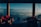 A luxurious seating area with wicker furniture and red cushions is set against a stunning backdrop of mountain ranges visible through large glass windows. The atmosphere is serene and inviting, providing a perfect blend of interior comfort with natural beauty. The misty layers of mountains stretch into the distance under a clear blue sky.