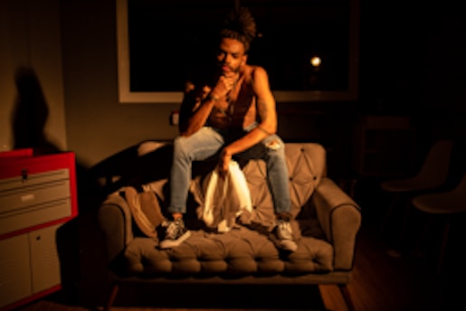 A person sits casually on a tufted sofa in a dimly lit room, wearing casual clothing, including a sleeveless shirt and ripped jeans. The lighting casts shadows and highlights the person's thoughtful pose and expression. The room contains minimal furniture, including a red cabinet on the left, and additional chairs in the background.