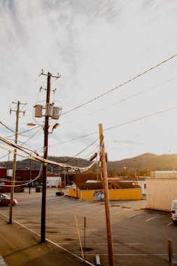 A view of a parking lot with several utility poles and power lines in the foreground. The background features a series of buildings, including a red-brick structure and a yellow-painted wall, set against a backdrop of rolling hills. The scene appears to be captured during sunset, with warm light casting long shadows.