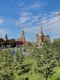 A landscape view featuring iconic Russian architecture with vibrant onion domes and tall spires. The foreground is full of lush green trees, adding contrast to the red brickwork of the historic buildings. The sky is clear with wisps of white clouds, giving a bright and open feel to the scene.