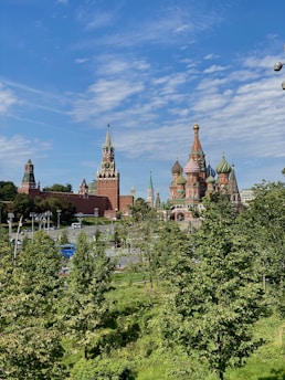 A landscape view featuring iconic Russian architecture with vibrant onion domes and tall spires. The foreground is full of lush green trees, adding contrast to the red brickwork of the historic buildings. The sky is clear with wisps of white clouds, giving a bright and open feel to the scene.