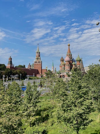 A landscape view featuring iconic Russian architecture with vibrant onion domes and tall spires. The foreground is full of lush green trees, adding contrast to the red brickwork of the historic buildings. The sky is clear with wisps of white clouds, giving a bright and open feel to the scene.